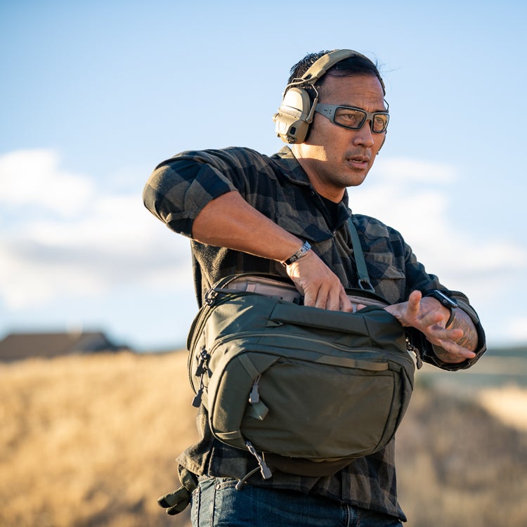 Photograph of a man in a flannel shirt and protective eyewear accessing his olive-green backpack in a rural setting with a distant view of houses under a clear sky