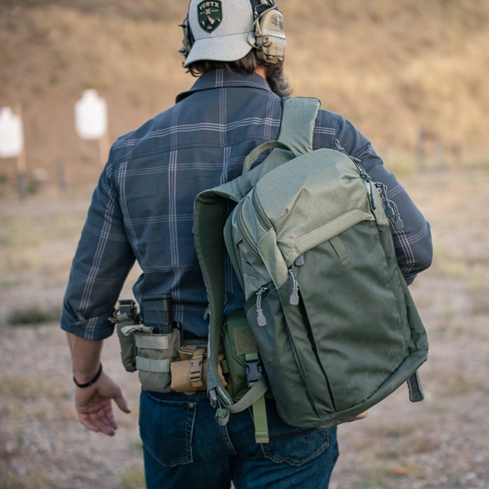 Photograph man wearing a Vertx backpack at an outdoor shooting range featuring target stands olive drab green backpack and a gray plaid shirt