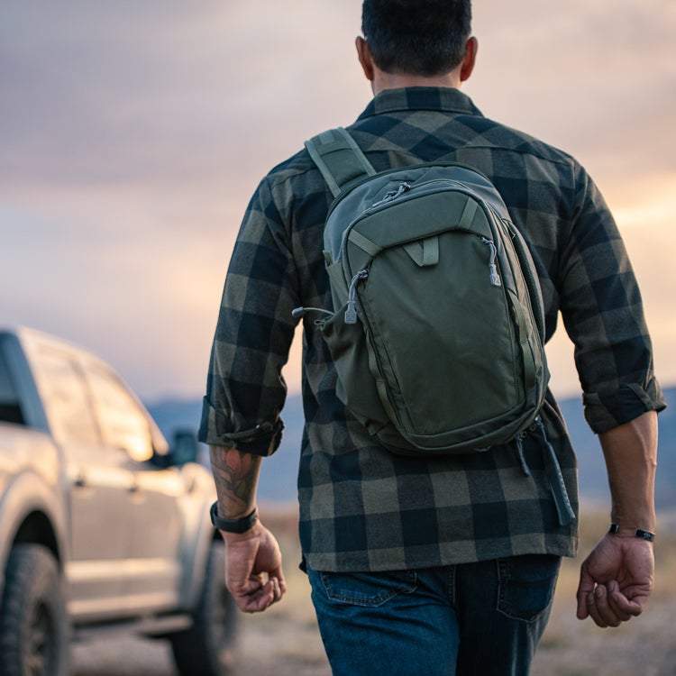 A photograph of a person wearing a green backpack and a dark green and black plaid shirt walking away from a silver truck in a desert landscape with a sunset sky featuring warm oranges and purples