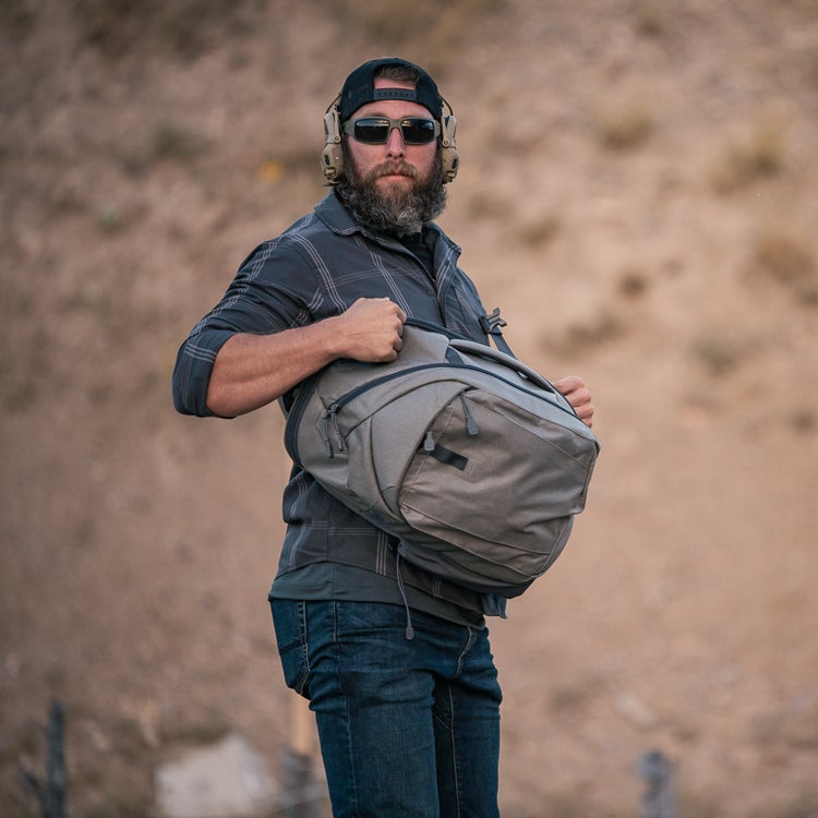 A photograph of a man with a beard wearing a plaid shirt and sunglasses at an outdoor shooting range carrying a large grey duffel bag with black accents and tan earmuffs.