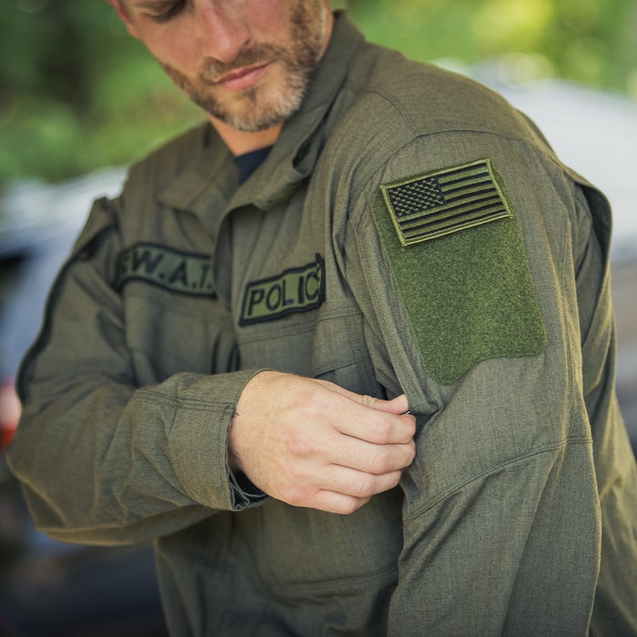 Photograph of a man adjusting his olive green SWAT police uniform featuring a US flag patch, black POLICE and SWAT lettering, and a textured fabric outdoors