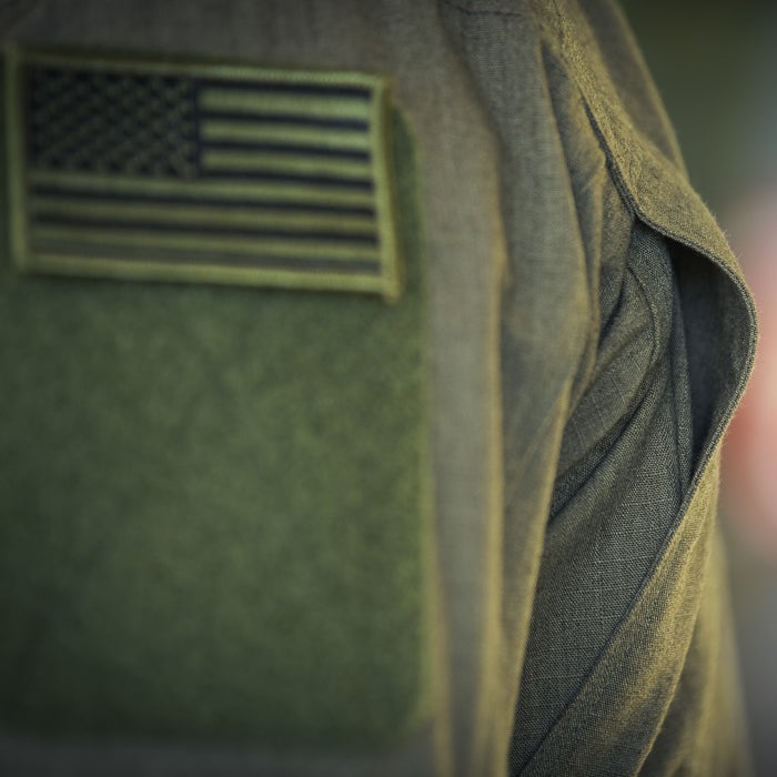 Close-up photograph of a military uniform detail featuring an olive green fabric texture a subdued US flag patch and subtle shadowing effects