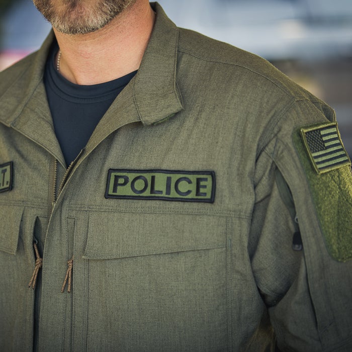 Close-up photograph of a police uniform featuring a POLICE patch, an American flag patch, and olive-green fabric with detailed stitching and drawstring pulls.