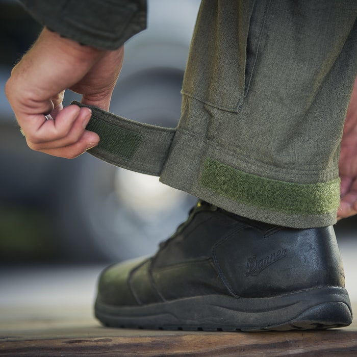 Close-up photograph showing a person adjusting the olive green fabric and Velcro straps of tactical pants with black Danner boots the fabric texture is visible along with the dark brown wooden surface