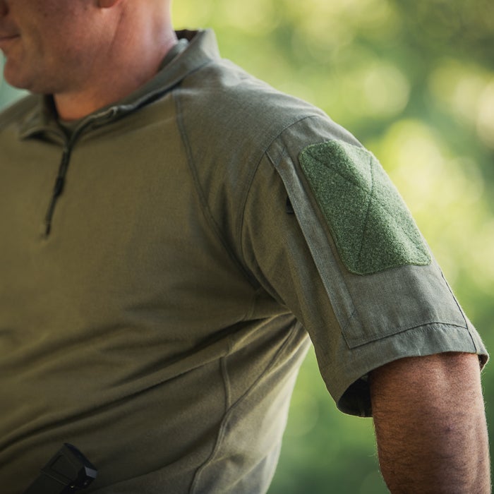 Photograph showing a close-up of a man's olive-green tactical short-sleeve shirt with a dark-green velcro patch on the sleeve and a hint of a firearm in the pocket against a blurred green natural background