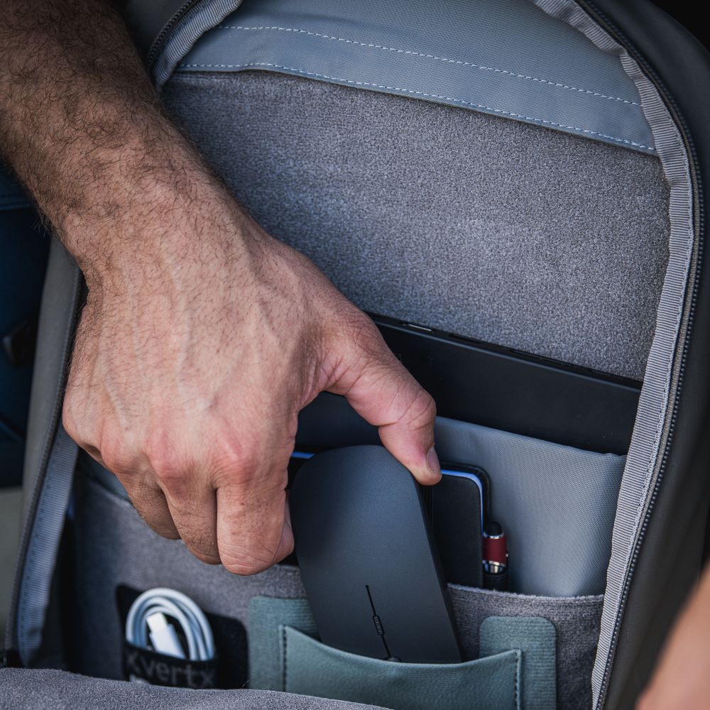 Photograph showing a man's hand placing a dark gray wireless mouse into an organized gray backpack pocket containing a tablet, notebook, and USB cable alongside other tech accessories