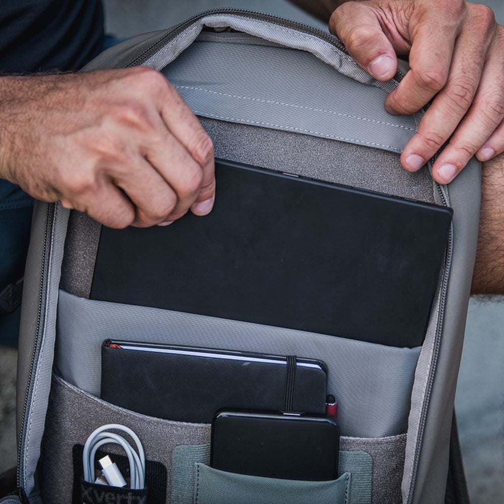 Photograph showing a person packing a gray backpack with a tablet notebook phone and USB cable inside the main compartment
