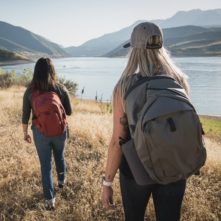 Photograph two women with backpacks hiking in a dry, golden field overlooking a lake and mountains with a red and grey backpack a stylish baseball cap and unique arm tattoos