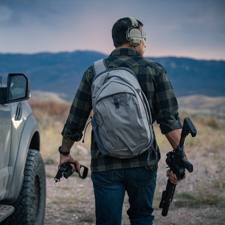 Photograph man carrying a grey backpack and rifle in a mountainous landscape near a silver truck featuring a muted sunset sky and dark green vegetation
