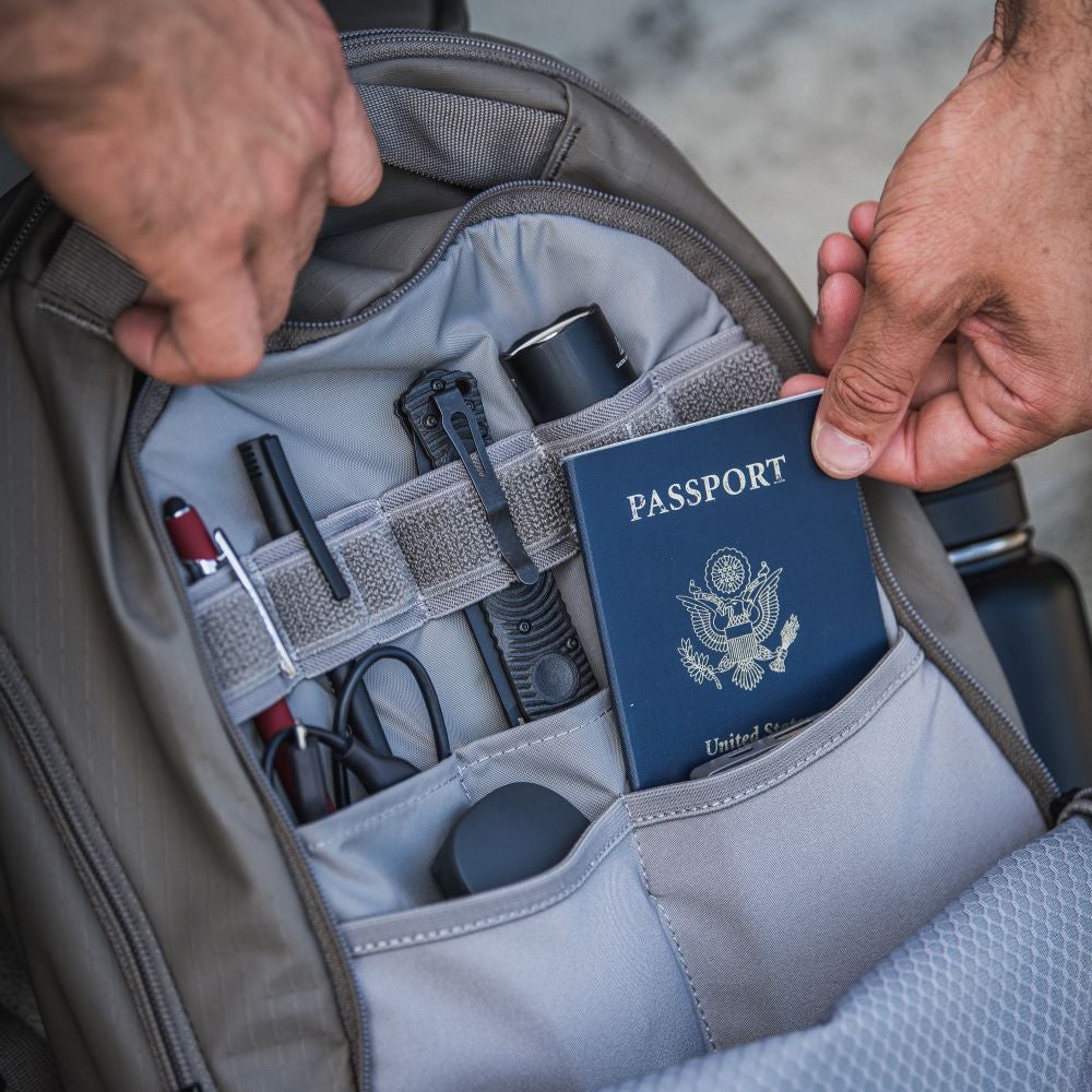 Photograph showing a grey backpack's organized interior containing a US passport, black tactical knives, a flashlight, and other travel essentials in a meticulously arranged setup