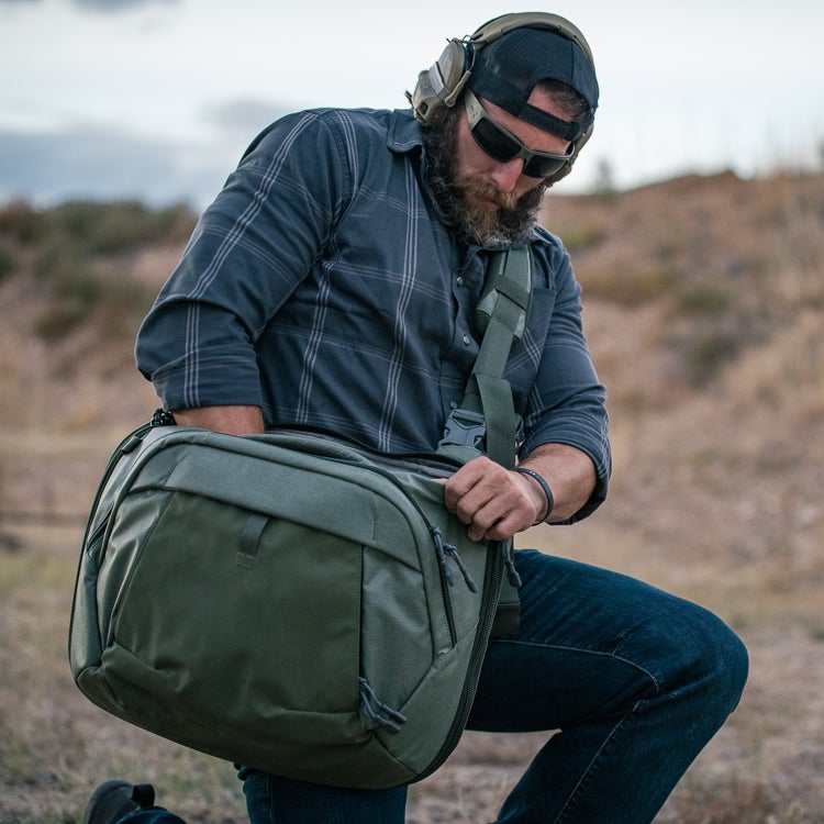 Photograph of a man in a dark plaid shirt and jeans accessing a large olive-green sling bag outdoors against a desert backdrop featuring muted earth tones and a tan hillside