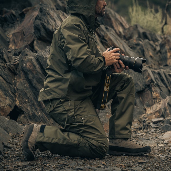Photograph of a person kneeling amidst dark rocks and using a Nikon D850 camera wearing olive green clothing and sturdy brown boots near a quarry.