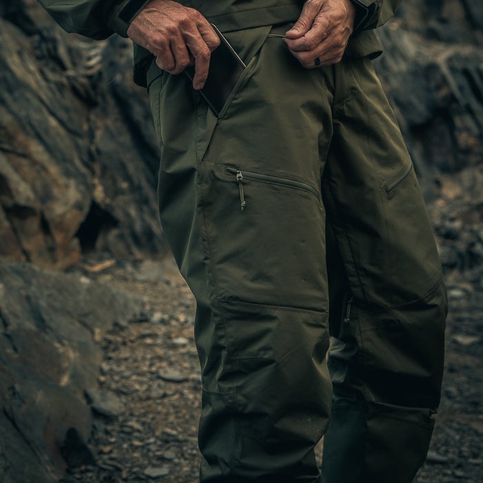Photograph of olive green outdoor pants with multiple pockets a person is placing a phone into a pocket against a rocky mountain backdrop featuring dark gray and brown stones