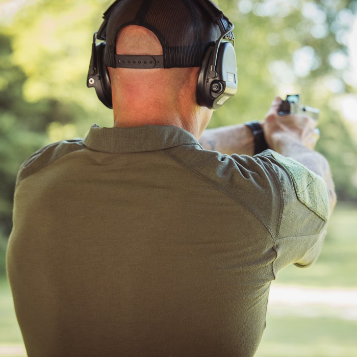 Photograph of a man wearing a green shirt and black hearing protection firing a handgun at an outdoor shooting range featuring leafy green trees and a partially visible olive-green tactical patch