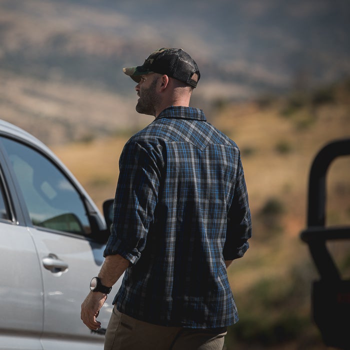 Photograph man wearing blue and gray plaid shirt and camouflage hat walking near a silver car against a mountainous backdrop featuring tan hills and a partly visible dark vehicle