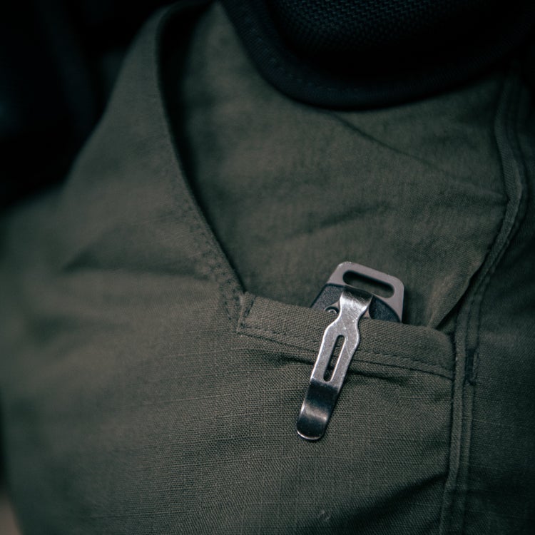 Close-up photograph of a metallic pocket knife with a unique clip in a dark olive green shirt pocket showing subtle textural details and muted color tones