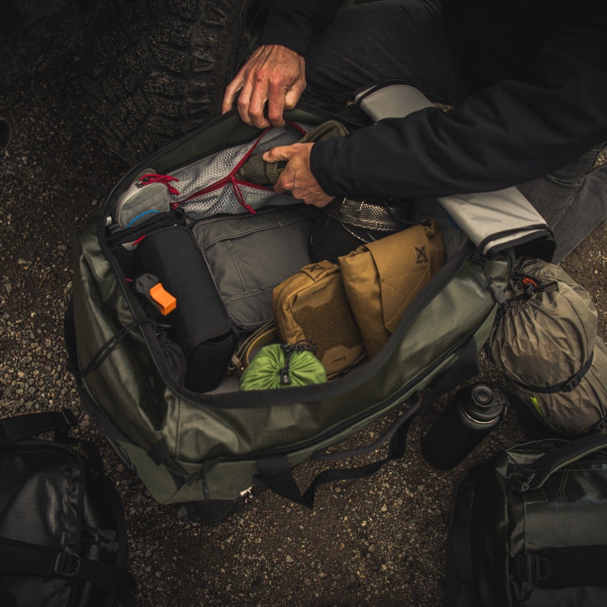 Photography A person packs a green duffel bag outdoors containing hiking boots, pouches, and a water bottle near other bags on gravel