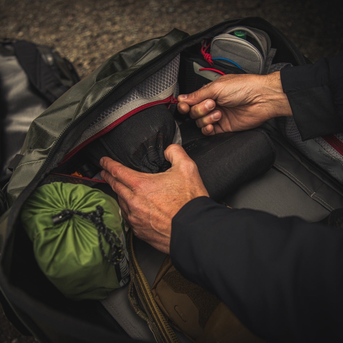 Photograph showing hands packing a dark-green backpack with red zippers containing olive-green stuff sack black mesh pouches and tan accessories