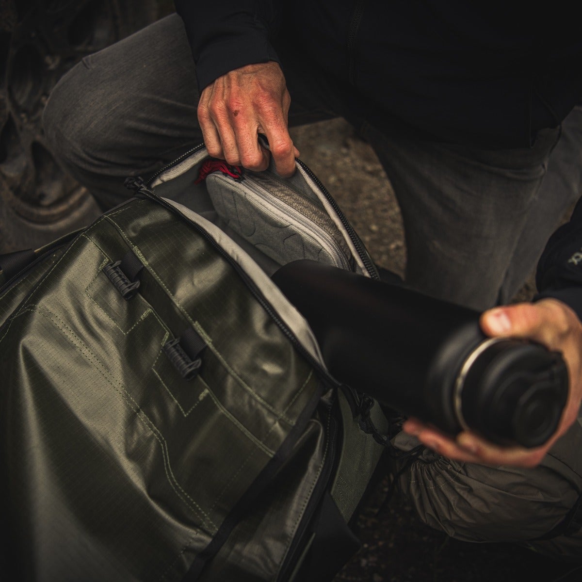 A photograph shows a person packing a dark-green, waterproof duffel bag with multiple zippered compartments, a gray inner pouch, and a black water bottle featuring a silver lid
