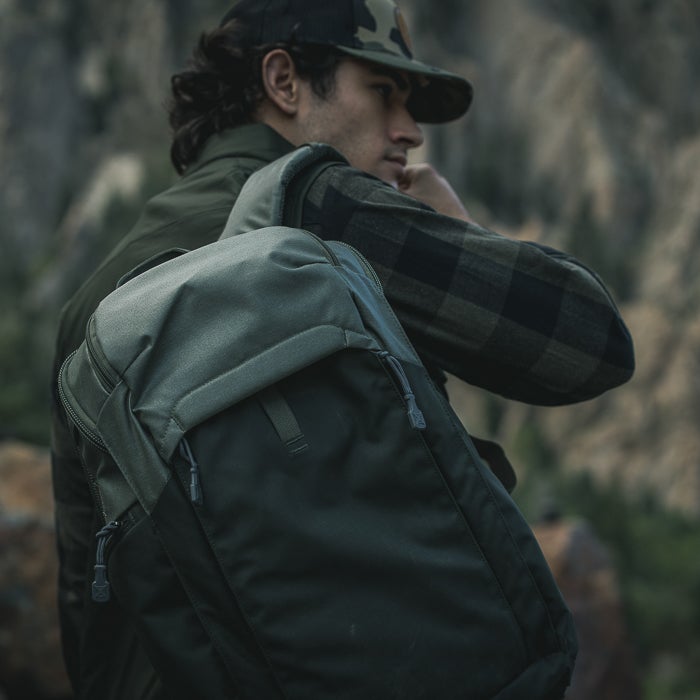 Photograph of a man wearing a dark green backpack with grey accents and a flannel shirt in a mountainous area with dark rock formations