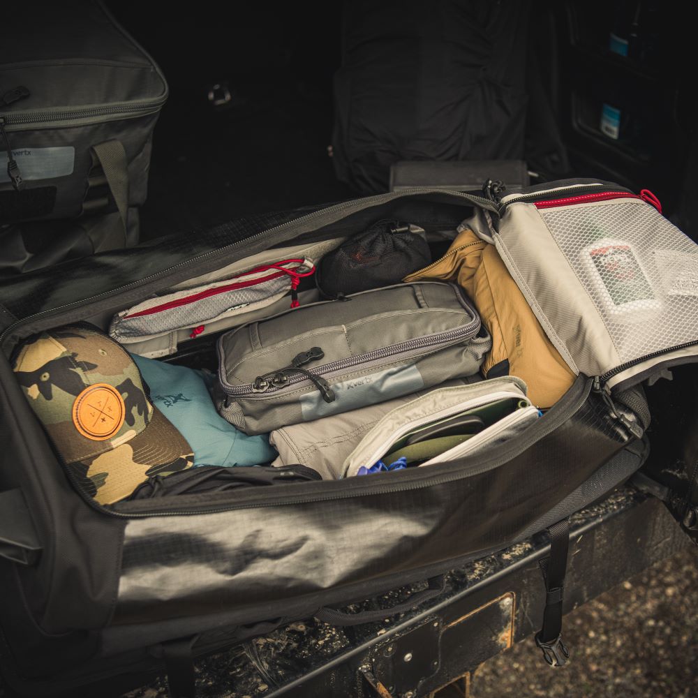 Photograph of a packed duffel bag in a vehicle trunk showing a camo hat gray and tan pouches and red zipper pulls
