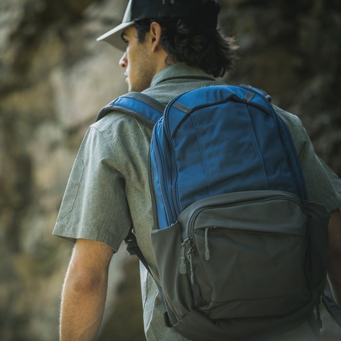 Photograph man wearing blue and gray backpack hiking near rocky cliffs showcasing durable zippers and adjustable straps