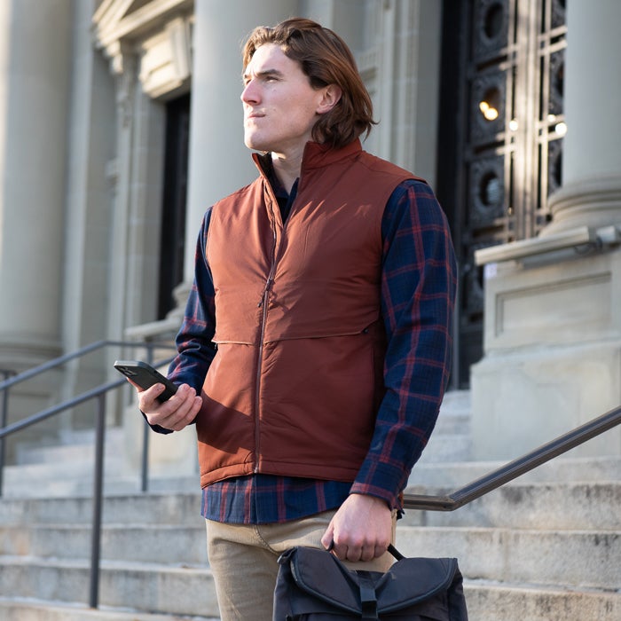 Photograph a man in a rust-colored vest and plaid shirt standing on the steps of a classical building with ornate columns and a detailed doorway holding a smartphone and messenger bag