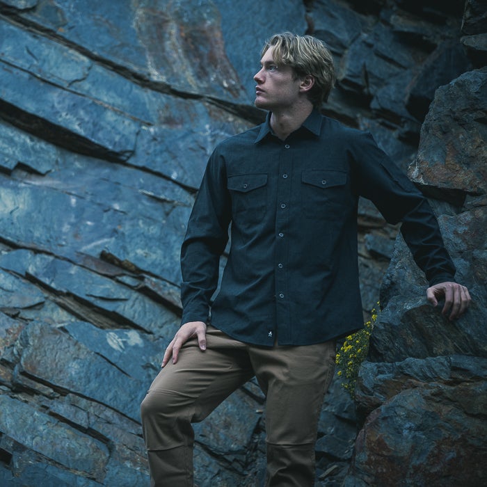 Photography Model wearing a dark shirt and tan pants standing against a dramatic slate rock face with hints of yellow wildflowers and deep blue-grey tones