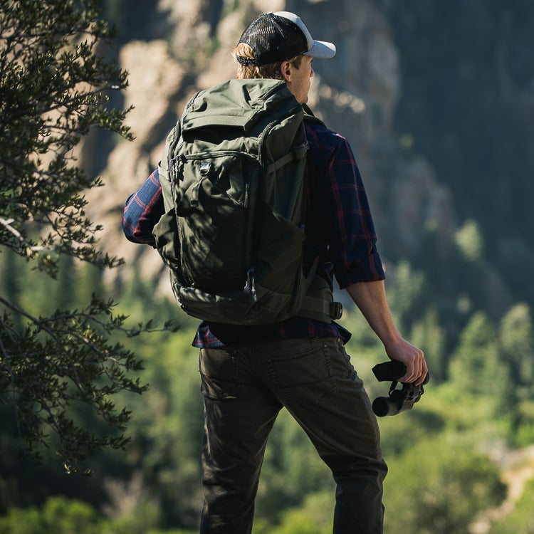 Photography A hiker with a large olive-green backpack and a red and black plaid shirt observes a mountainous landscape featuring rugged cliffs and lush greenery while holding binoculars in his hand