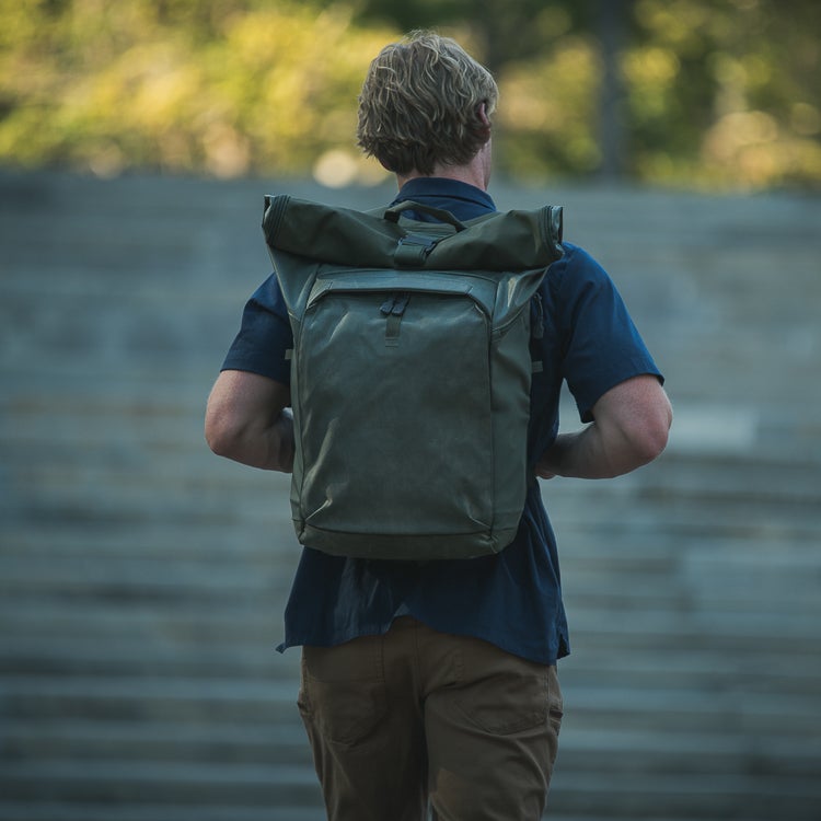 Photograph of a man with blond hair wearing a dark-olive roll-top backpack near a stone staircase showing muted greens and blues with a hint of sunlit background