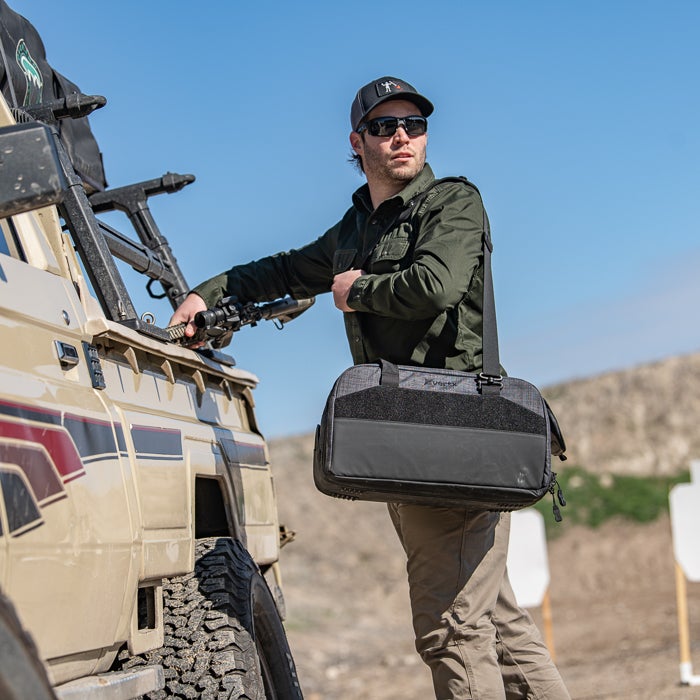 Photograph man with rifle near a vintage Toyota Land Cruiser at an outdoor shooting range featuring tan pants, a dark green shirt, and a black gun bag with MOLLE webbing