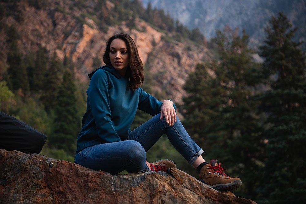 Photography Woman in teal hoodie and blue jeans sits on a rock overlooking a mountain range with dark green pine trees and reddish-brown rock formations wearing brown hiking boots with red laces