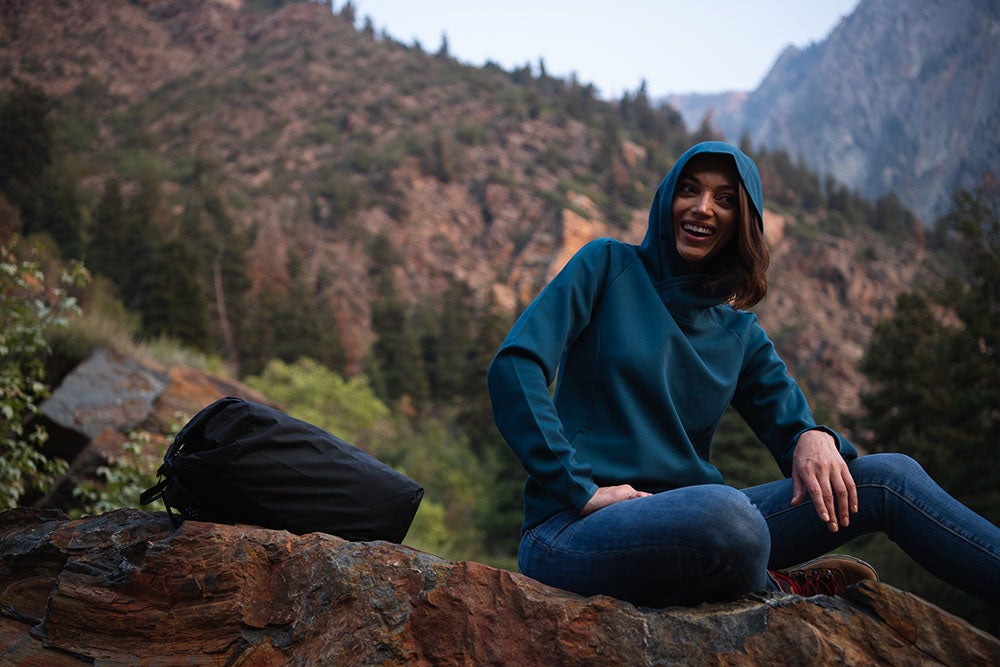 Photograph of a woman in a teal hoodie and blue jeans sitting on a rock in a mountainous landscape with lush green vegetation and dark brown rocky cliffs a black backpack nearby