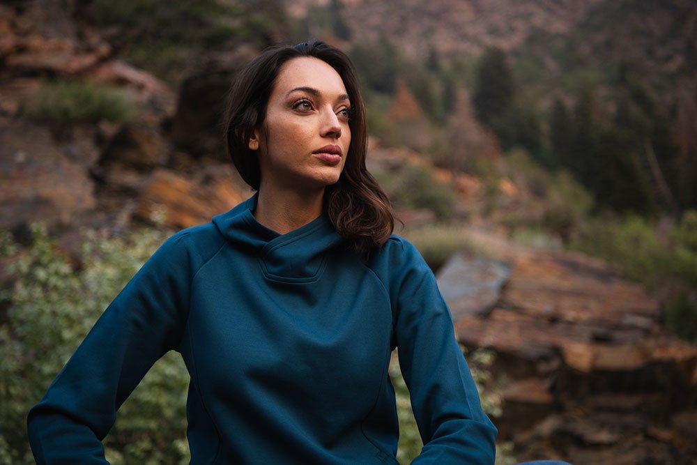 Photography of a woman in a teal hoodie seated outdoors amongst rocks and trees a mountainous backdrop showing varied textures and earthy tones