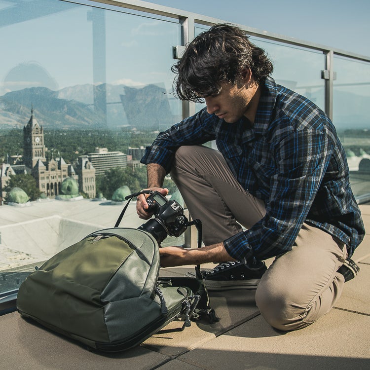 Photograph of a man in a blue plaid shirt and khaki pants kneeling on a rooftop in Salt Lake City Utah placing a camera into a green backpack with the Utah State Capitol building and Wasatch Mountains visible in the background showcasing the photographer's olive-toned backpack and the camera's lens