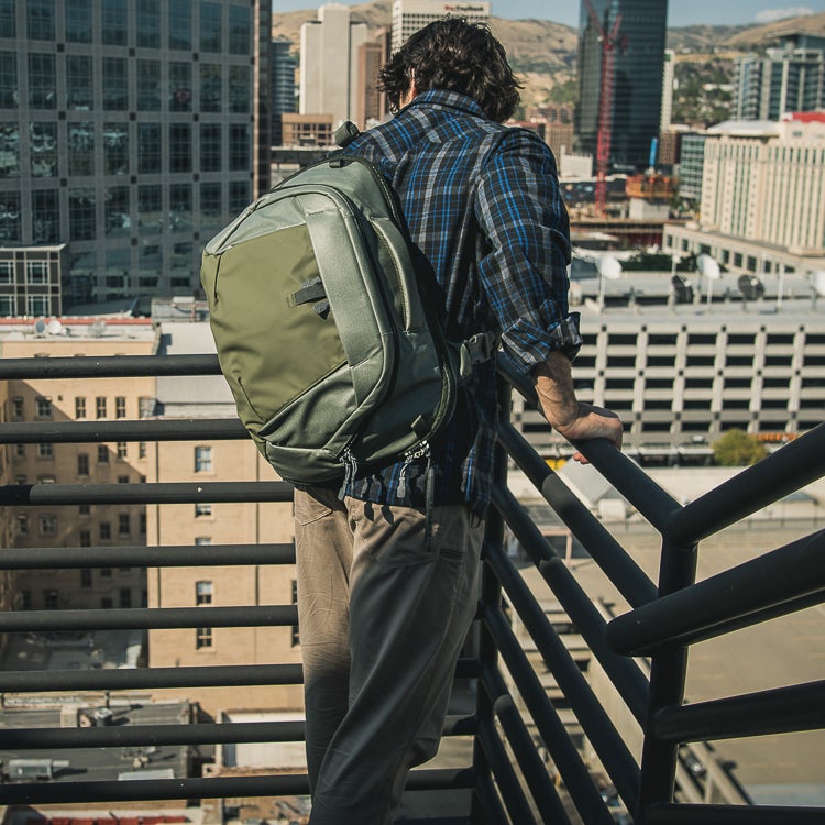 Photograph of a man with a large olive-green backpack leaning on a rooftop railing overlooking a city featuring a multi-story parking garage and distinctive buildings showcasing blue sky and vibrant sunlight