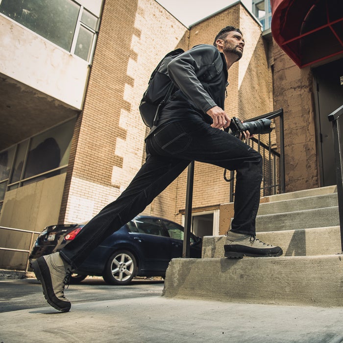 Photograph of a man with a backpack and camera ascending urban steps near brick buildings and a parked car featuring grey hiking boots and a dark jacket
