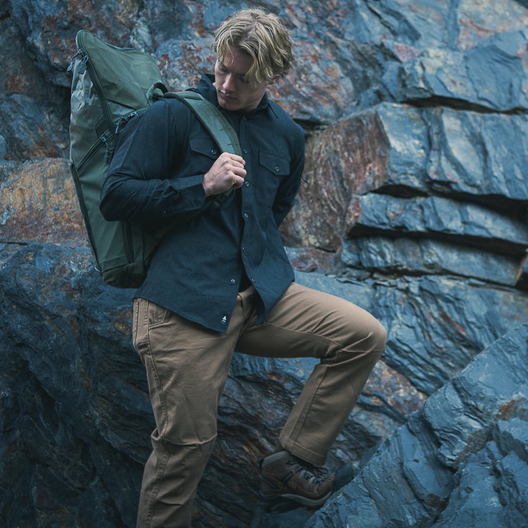 Photograph model wearing dark shirt tan pants and large olive green backpack against a rocky mountain backdrop featuring layered slate blue and brown stones