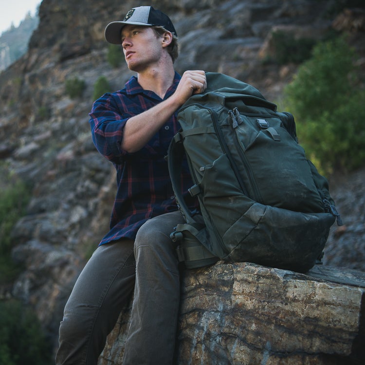 Photograph of a man with a large olive-green backpack sitting on a rock in a mountainous area featuring rugged cliffs and a red and blue plaid shirt