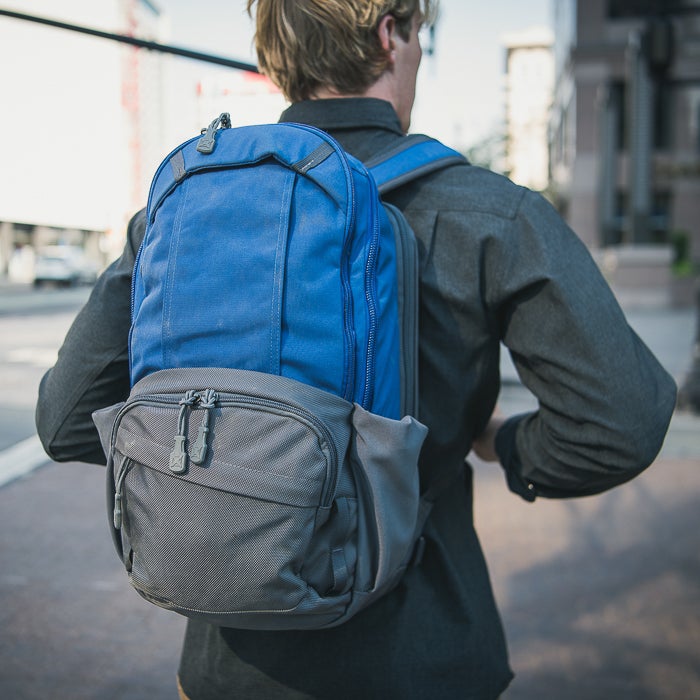 Photograph of a person wearing a blue and gray backpack with unique zipper pulls on a city street featuring a modern building and a traffic light