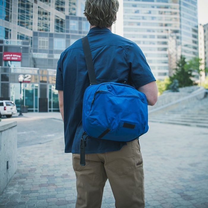 Photograph man with vibrant blue crossbody bag walking in city with modern architecture and green trees a lamppost and a building with Apple Store signage visible