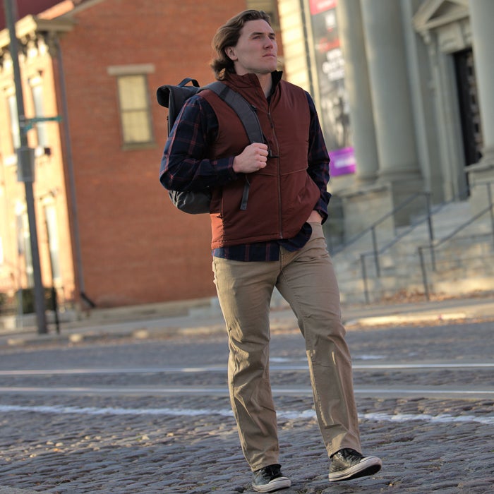 Photograph of a man in a maroon vest and khaki pants walking down a cobblestone street past brick buildings and classic columns a gray backpack and plaid shirt are also visible