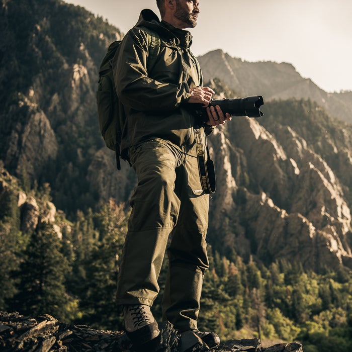 Photograph of a man in olive green hiking attire holding a camera with a Nikon strap amidst a mountainous landscape featuring rugged cliffs and verdant pine trees brown rocky outcropping and warm sunlight