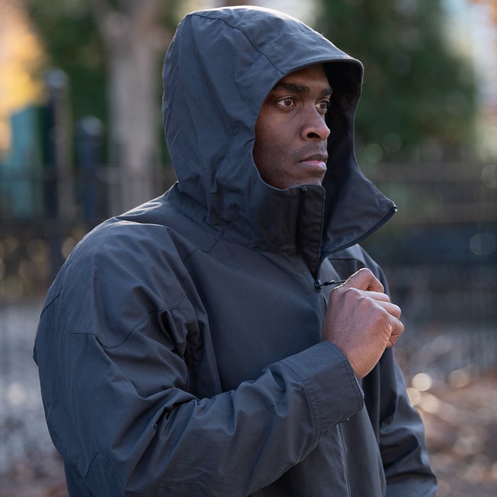 Photograph of a man in a dark hooded jacket outdoors near a fence and blurred trees showing his serious expression and the jacket's details like its zipper and texture