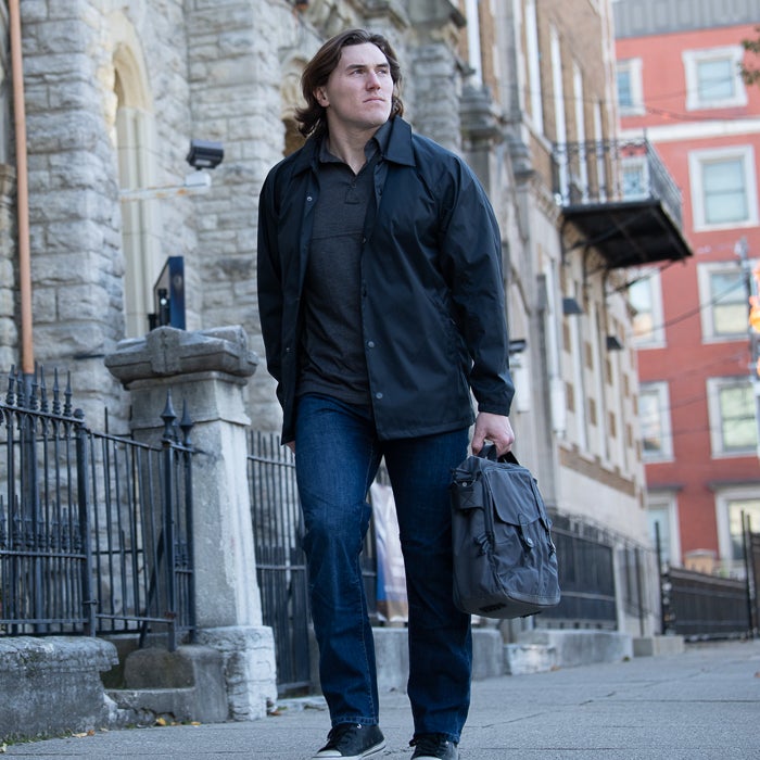 Photography Man walking down a city street past a stone church with wrought iron fence and a red brick building carrying a grey messenger bag his dark jacket and blue jeans stand out against the urban background