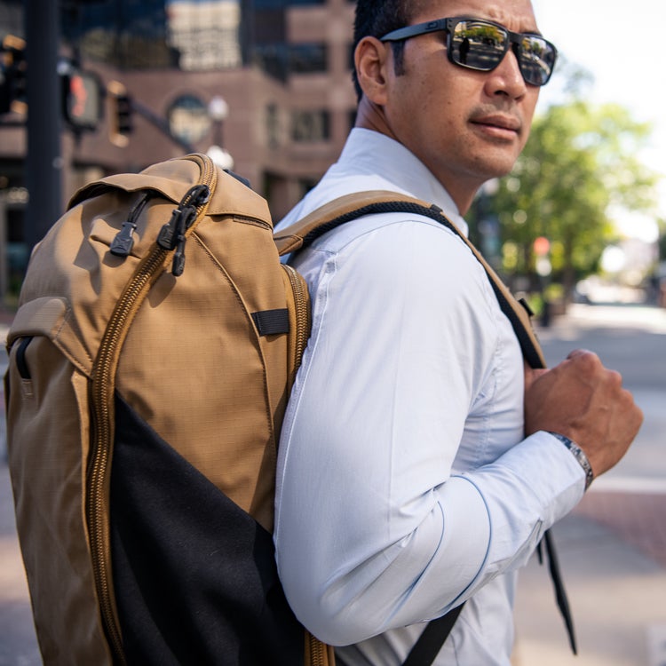 Photograph of a man in a light blue shirt and sunglasses carrying a tan and black backpack with multiple zipper pulls in an urban setting featuring modern buildings and traffic lights