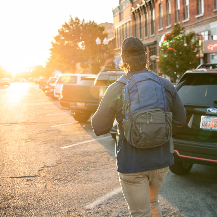 Photograph person walking away with blue backpack down a sunlit street lined with parked cars near historic brick buildings with warm sunset light lens flare and vibrant autumn colors