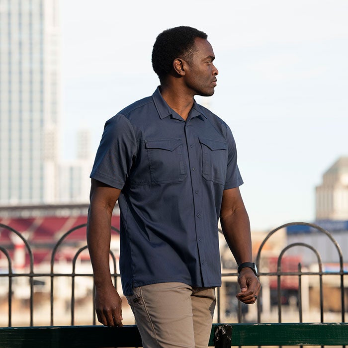 Photography model wearing a dark blue short-sleeved shirt and khaki pants walks past a green bench and wrought iron fence with a blurred cityscape including skyscrapers and a stadium in the background