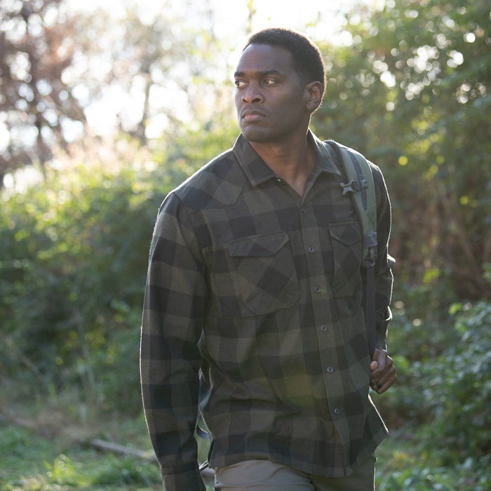 Photograph of a man wearing a dark green and black plaid shirt hiking through a sun-dappled forest with a backpack his serious expression and the warm light creating a mood of quiet contemplation