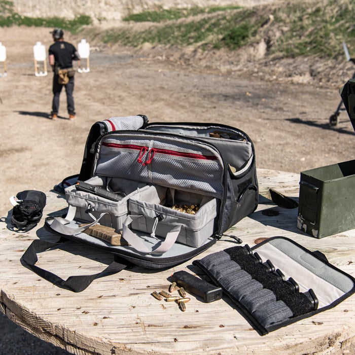 Photograph open range bag at an outdoor shooting range with targets in the background featuring gray fabric interior red zipper pulls and brass cartridges.